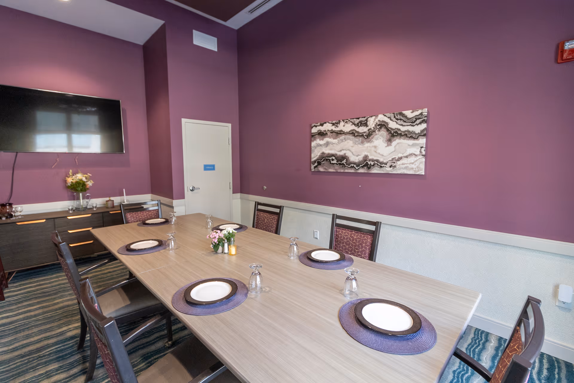 Dining room with a long table set with plates and glasses, purple walls, a mounted TV and abstract wall art.
