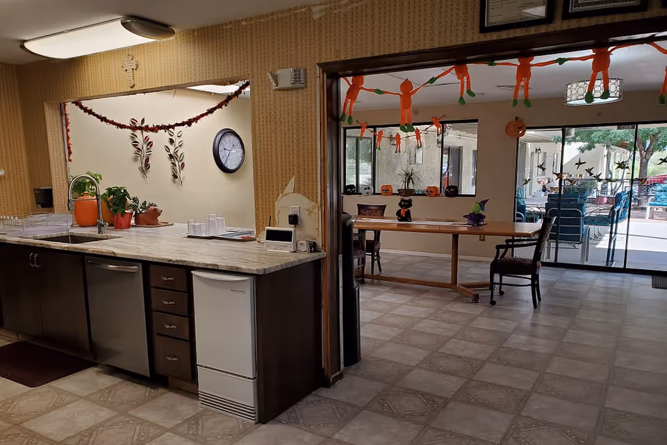 Open common area with a kitchenette counter in the foreground and a decorated dining/activity room with tables and sliding glass doors to an outdoor patio.