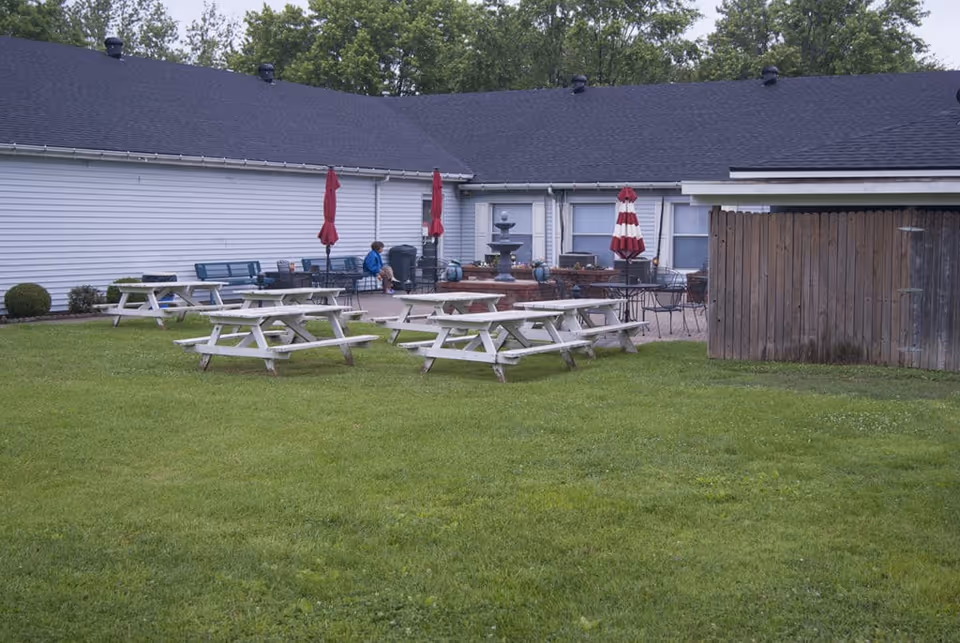 Outdoor area of a senior living facility with green grass, several white picnic tables, a patio with red umbrellas, a water fountain, and a person sitting on a bench near the building.