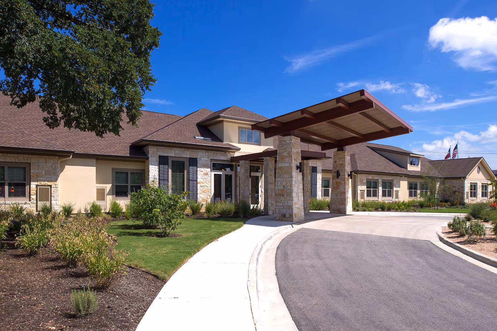 Exterior view of Morningside House of Sarasota, showing a single-story building with stone and beige walls, a covered entrance supported by stone pillars, a curved driveway, landscaped greenery, and a bright blue sky with a few clouds.