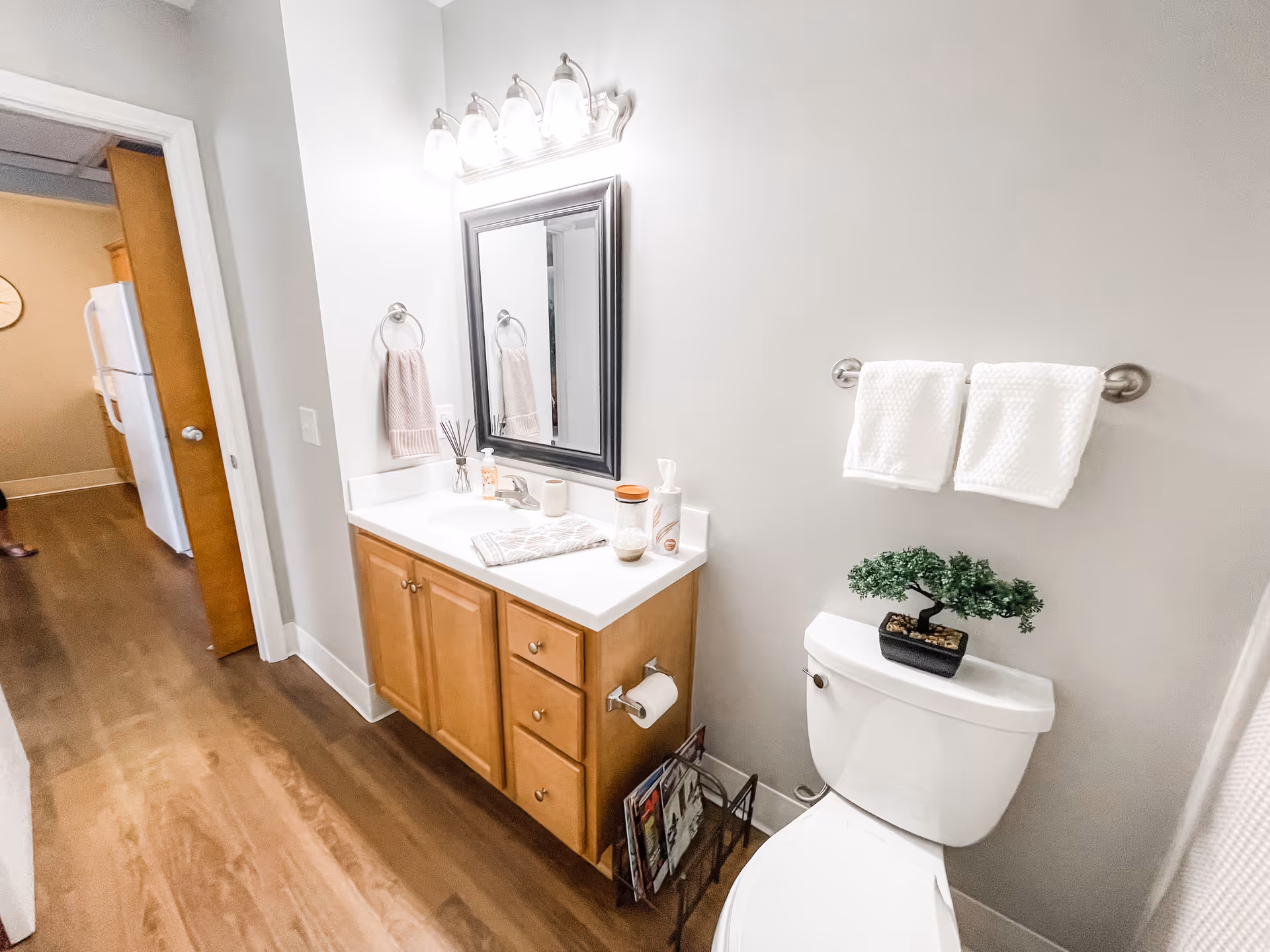 Bathroom with a wooden vanity and mirror, a toilet topped with a small potted plant, and towels on a wall rack.