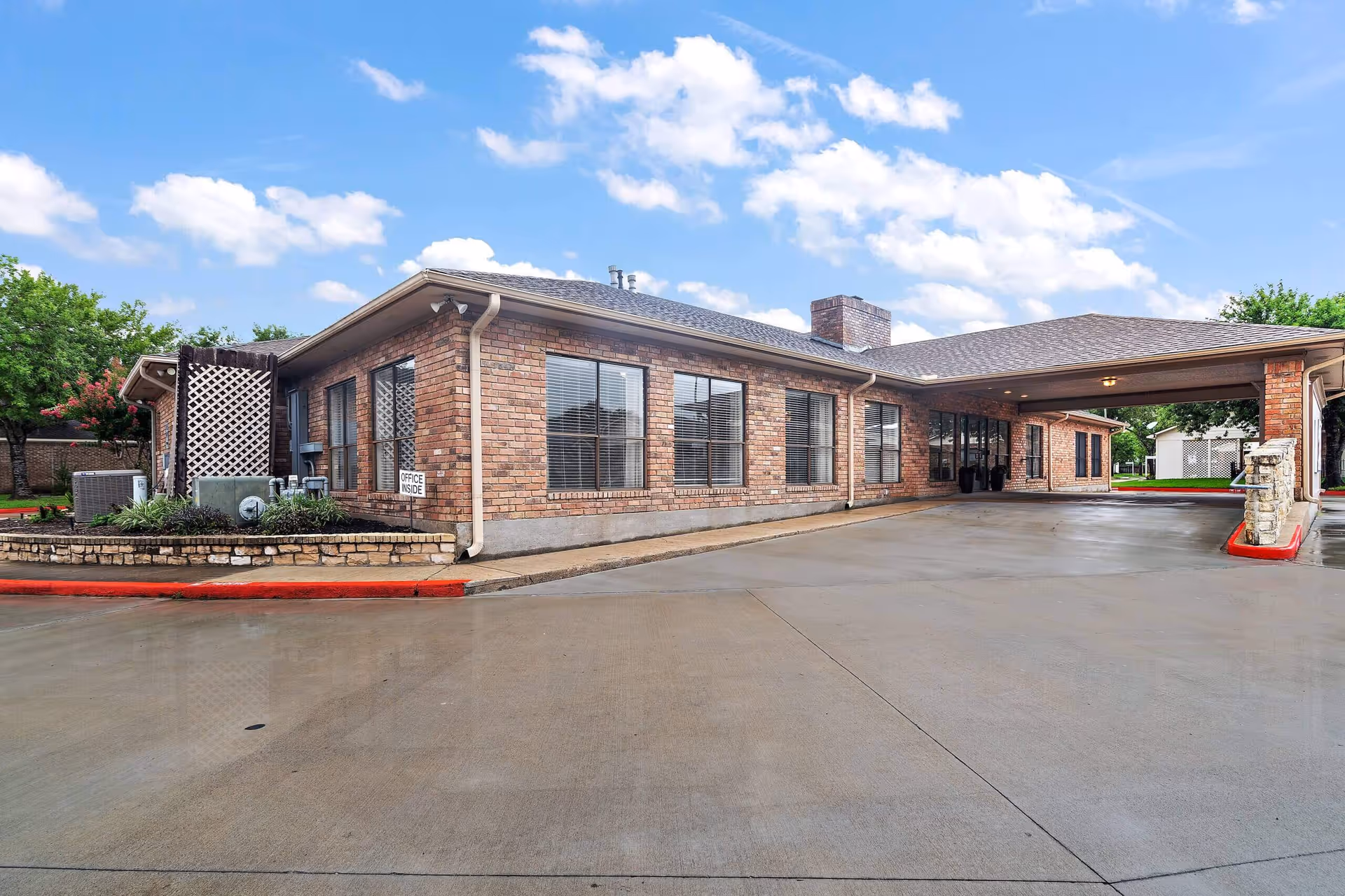 Exterior view of a single-story brick building with multiple windows and a covered entrance driveway. The building is surrounded by a concrete driveway and some greenery, with a partly cloudy blue sky in the background.
