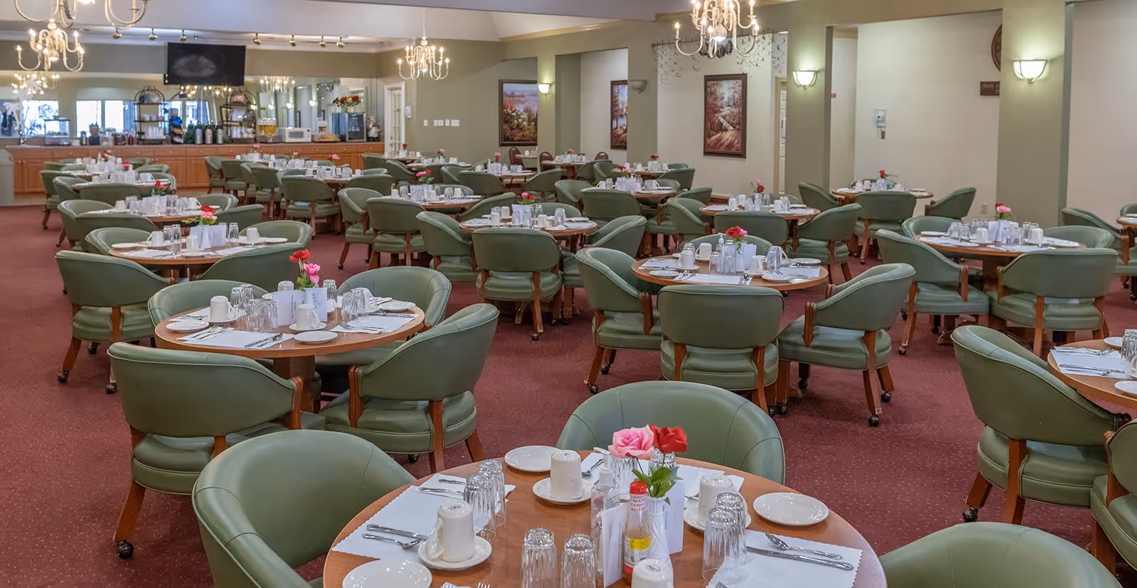 A spacious dining room with multiple round tables set for meals. Each table has green cushioned chairs, white plates, cups, silverware, glasses, and small flower arrangements. The room has chandeliers, wall sconces, framed artwork, and a buffet area with appliances and decorations in the background.