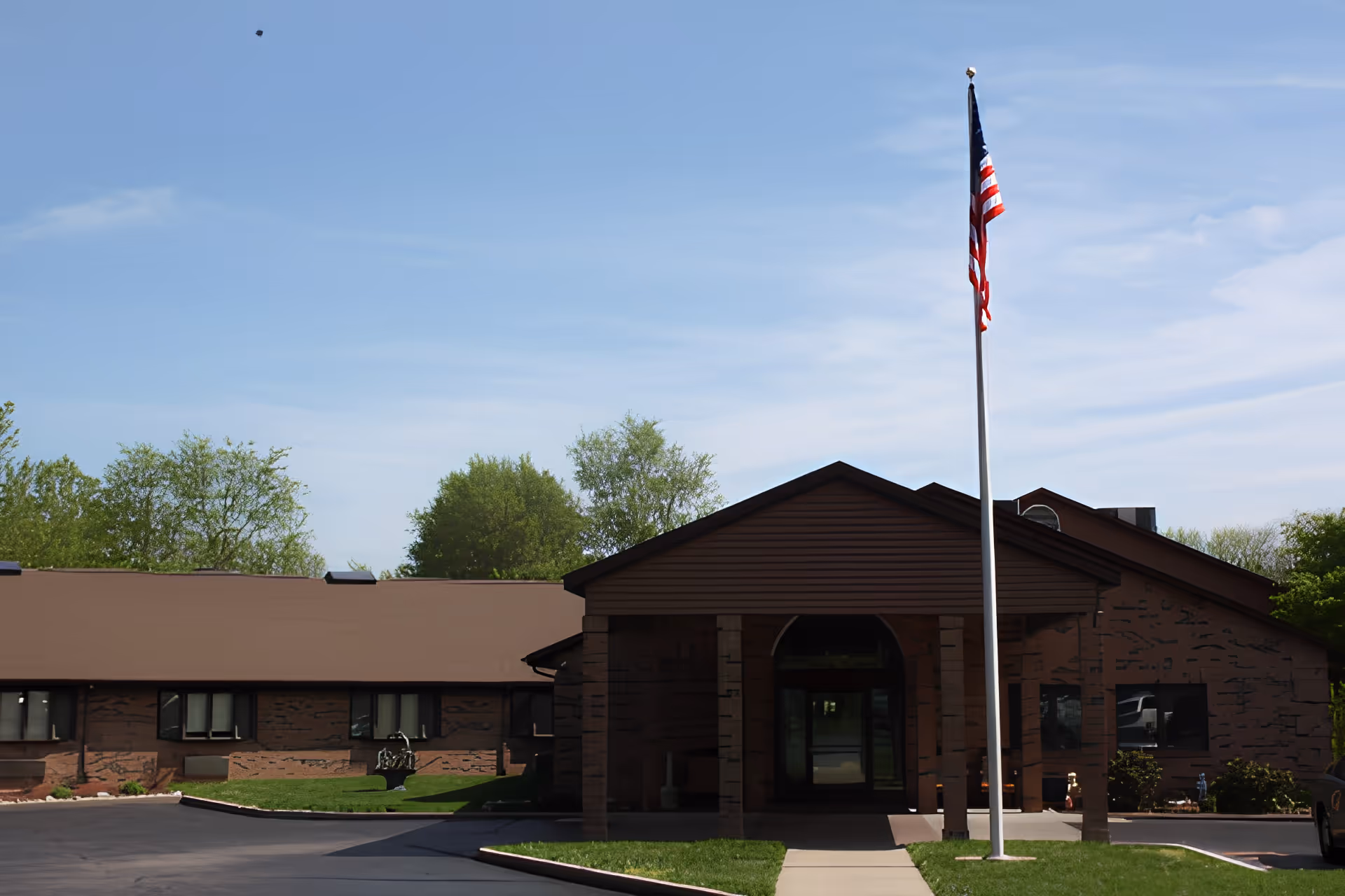 Single-story brick assisted living building entrance with a flagpole flying the American flag under a clear sky.