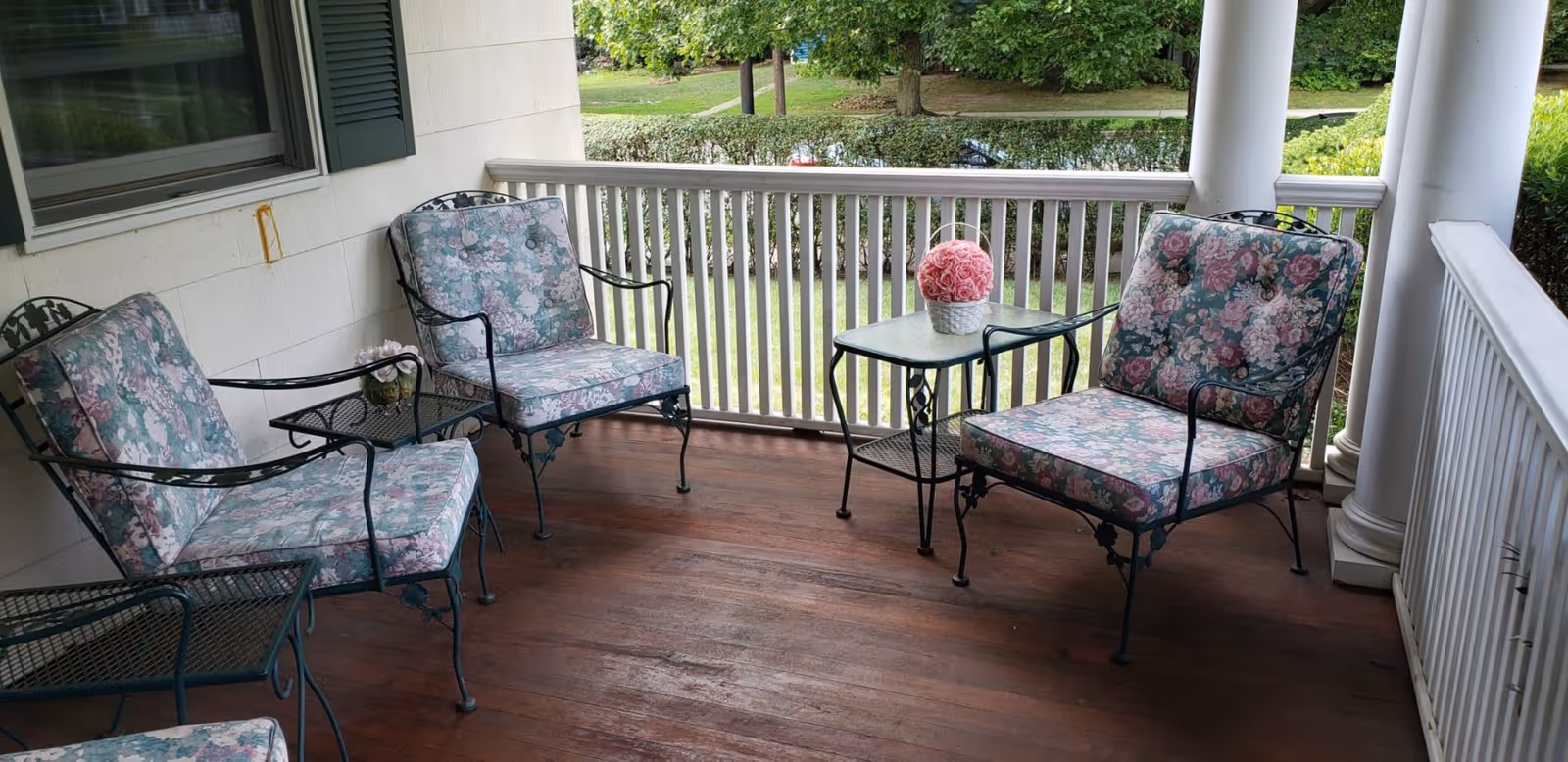 A covered porch with wooden flooring featuring three metal chairs with floral cushions and a small glass-top table holding a pink flower arrangement. The porch has white railings and columns, with green trees and grass visible in the background.