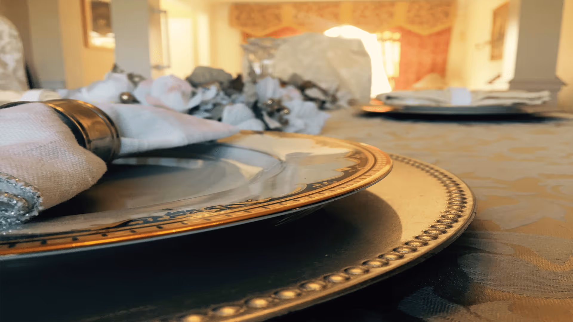 Close-up view of an elegant dining table setting with a decorative plate, a napkin with a napkin ring, and a tablecloth with a subtle pattern. The background shows a softly lit room with warm tones and blurred furniture.