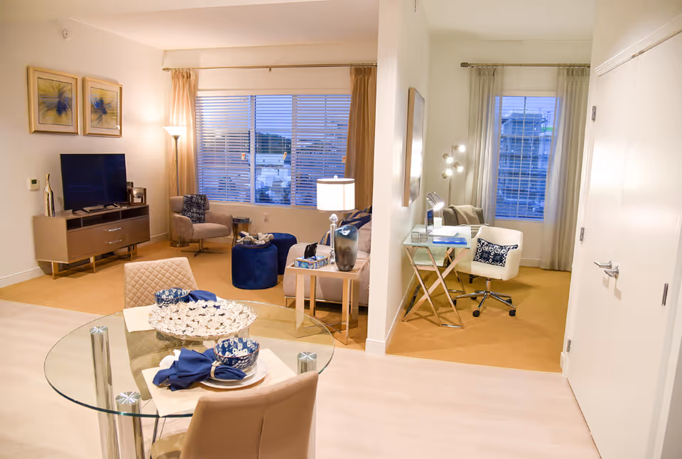 A bright and modern living space featuring a glass dining table set with blue and white dishware and beige chairs in the foreground. Beyond the dining area is a cozy living room with a TV on a stand, two armchairs, a sofa, and a side table with a lamp. To the right, there is a small office nook with a glass desk, a white swivel chair, and a floor lamp, all set against large windows with beige curtains.