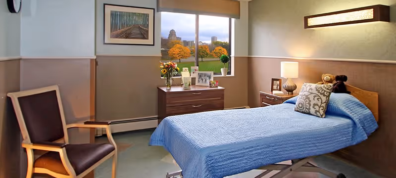 A cozy bedroom with a single bed covered in a blue quilt, a decorative pillow, and two teddy bears on the headboard. There is a wooden dresser under a window showing an outdoor view with trees and buildings. A wooden chair with dark cushions is placed near the bed. The room has soft lighting from a table lamp and a wall-mounted light fixture, with framed artwork on the wall.