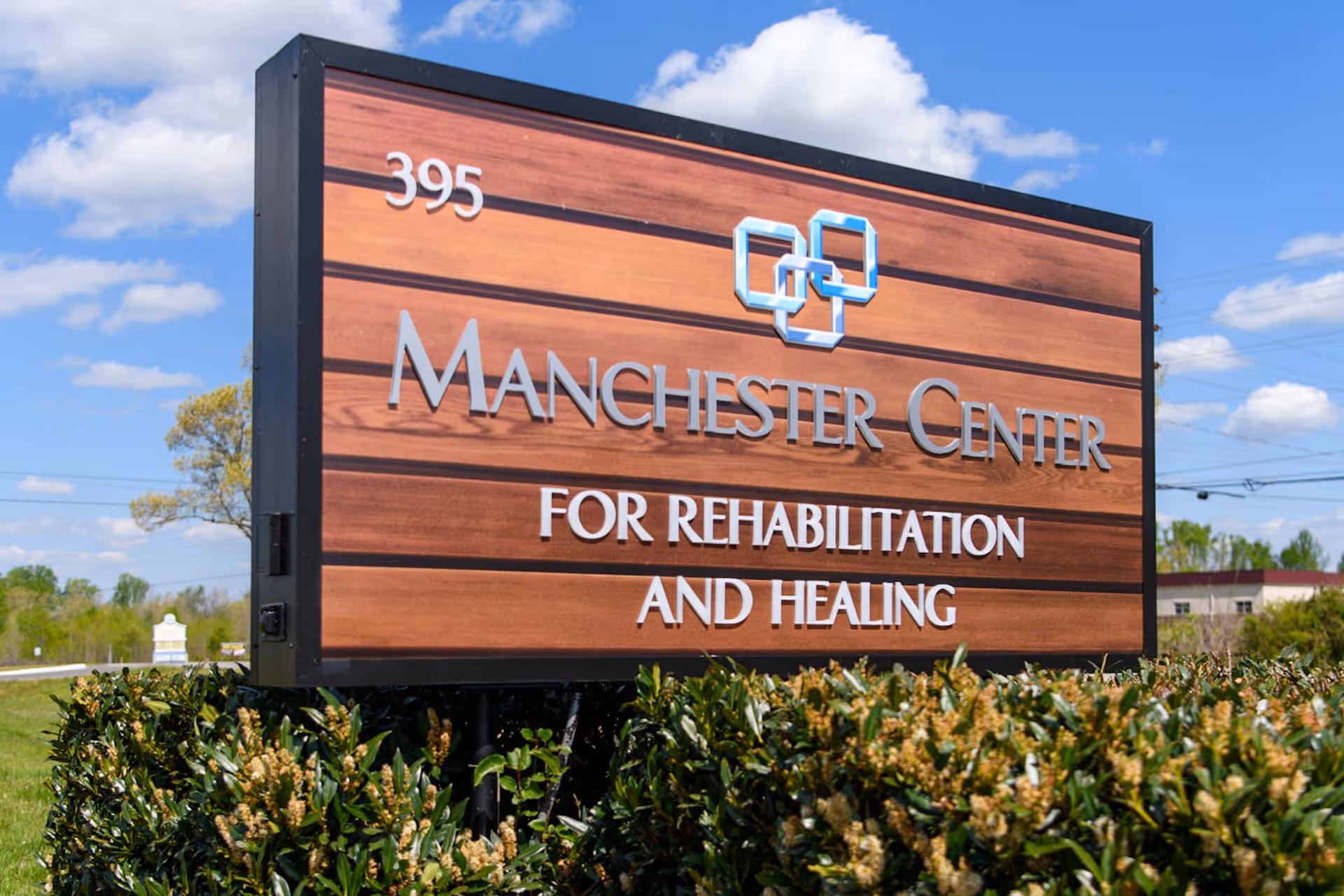 Outdoor wooden sign for Manchester Center for Rehabilitation and Healing with the number 395, surrounded by green bushes under a blue sky with some clouds.