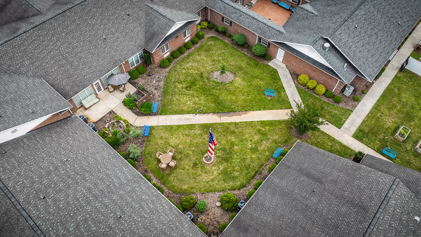 Aerial view of a grassy courtyard with intersecting concrete walkways, benches, patio furniture and an American flag, surrounded by brick buildings and rooftops.
