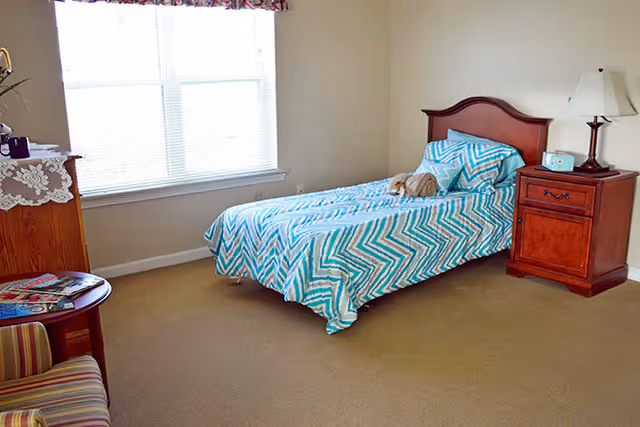 A simple bedroom with a single bed covered in a blue and white chevron patterned bedspread. There is a wooden nightstand with a lamp and a clock on it next to the bed. A window with blinds lets in natural light. A small round table with magazines and a striped upholstered chair are also visible.