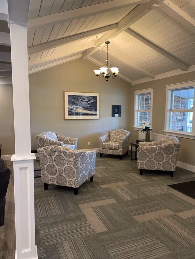 A cozy sitting area with four patterned armchairs arranged in a circle on a carpeted floor. The room has a vaulted ceiling with exposed beams painted white, a chandelier hanging from the ceiling, and two windows letting in natural light. A framed picture of a cloudy sky hangs on the beige wall, and a small table with a vase of flowers is placed near the windows.