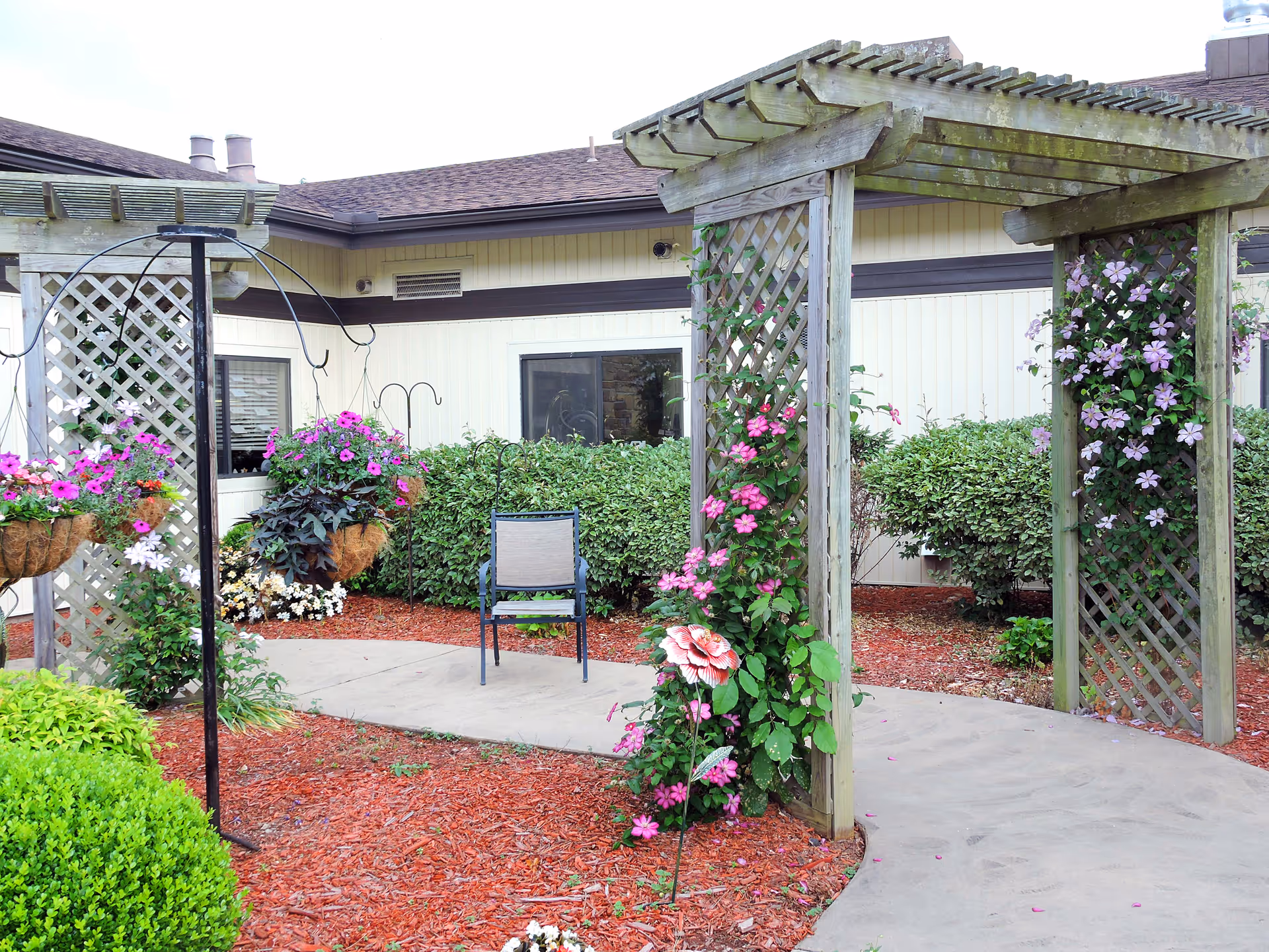 Outdoor garden area at Lake Forest Senior Living at Hot Springs Village featuring a wooden pergola with climbing pink and purple flowers, hanging flower baskets, a single chair on a curved concrete pathway, and well-maintained bushes and mulch beds.
