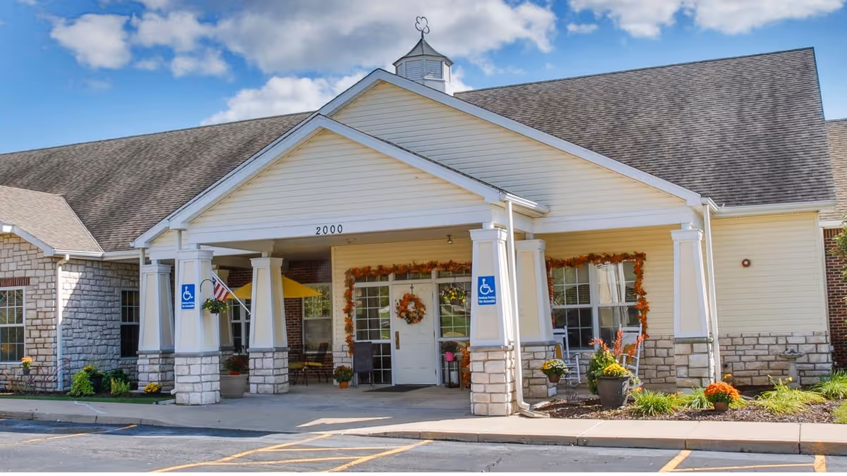 Front entrance of a single-story senior living facility with a covered portico, stone pillars, handicap signs, and autumn decorations.