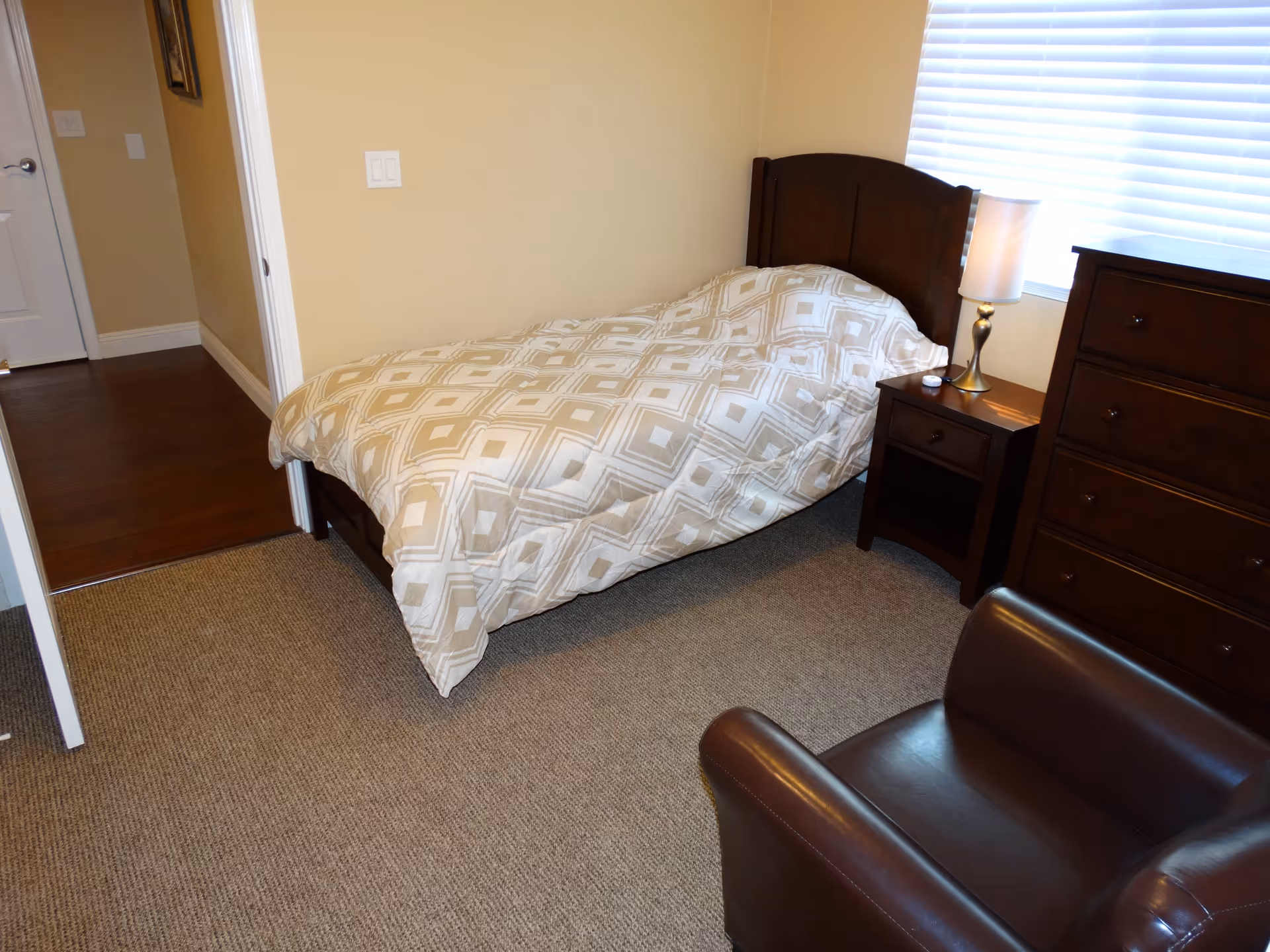 A small bedroom with a single bed covered with a beige and white patterned comforter. Next to the bed is a dark wooden nightstand with a lamp and a small item on it. There is a matching dark wooden dresser and a brown leather armchair in the room. The room has beige walls, a window with white blinds, and a carpeted floor.