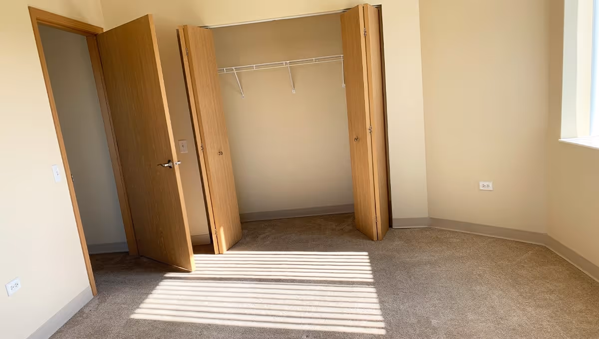 Empty carpeted bedroom with an open closet and wooden doors, sunlight casting blinds' shadows on the floor.