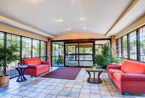 Bright entrance lobby of a senior living facility with large glass windows, two red couches facing each other, two small round tables with potted plants, tiled floor, and automatic sliding glass doors leading outside.