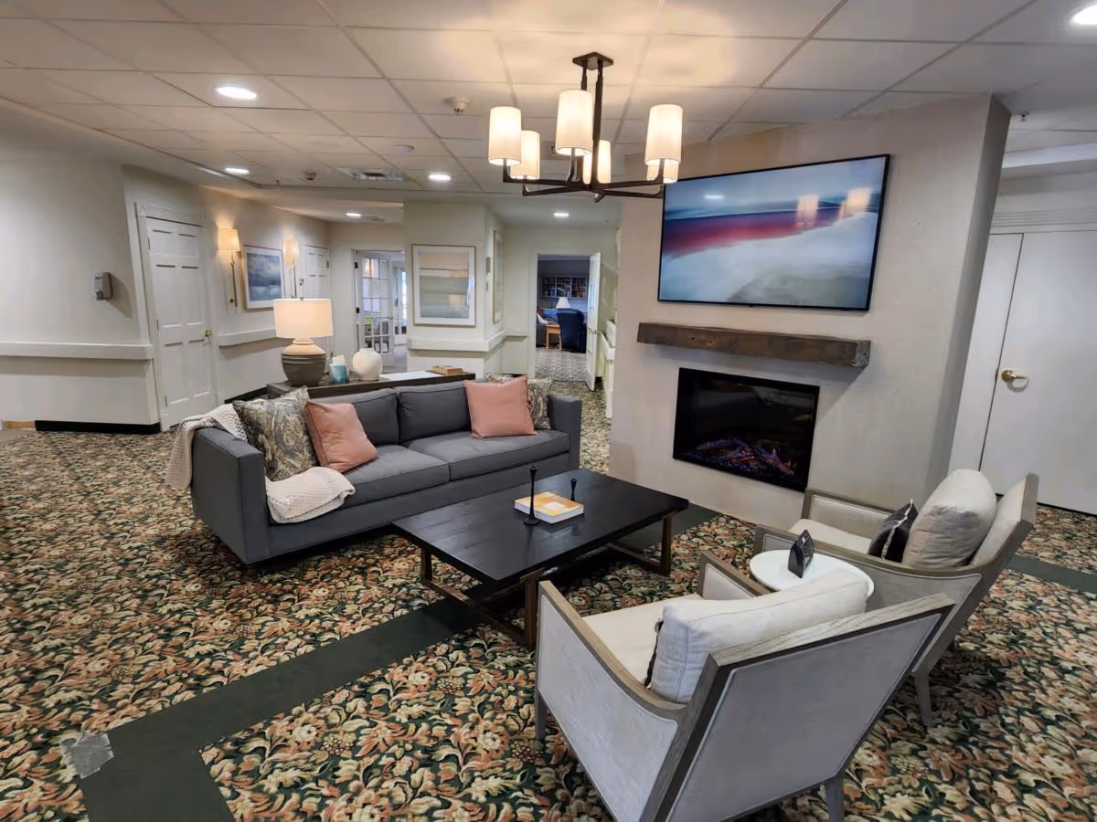 A cozy living room area in a senior living facility with a gray sofa adorned with pink and patterned pillows, two beige armchairs, and a dark wooden coffee table. The room features a patterned carpet, a modern chandelier, a wall-mounted flat-screen TV above a fireplace, and soft lighting from lamps and wall sconces. The background shows a hallway with doors and framed artwork on the walls.