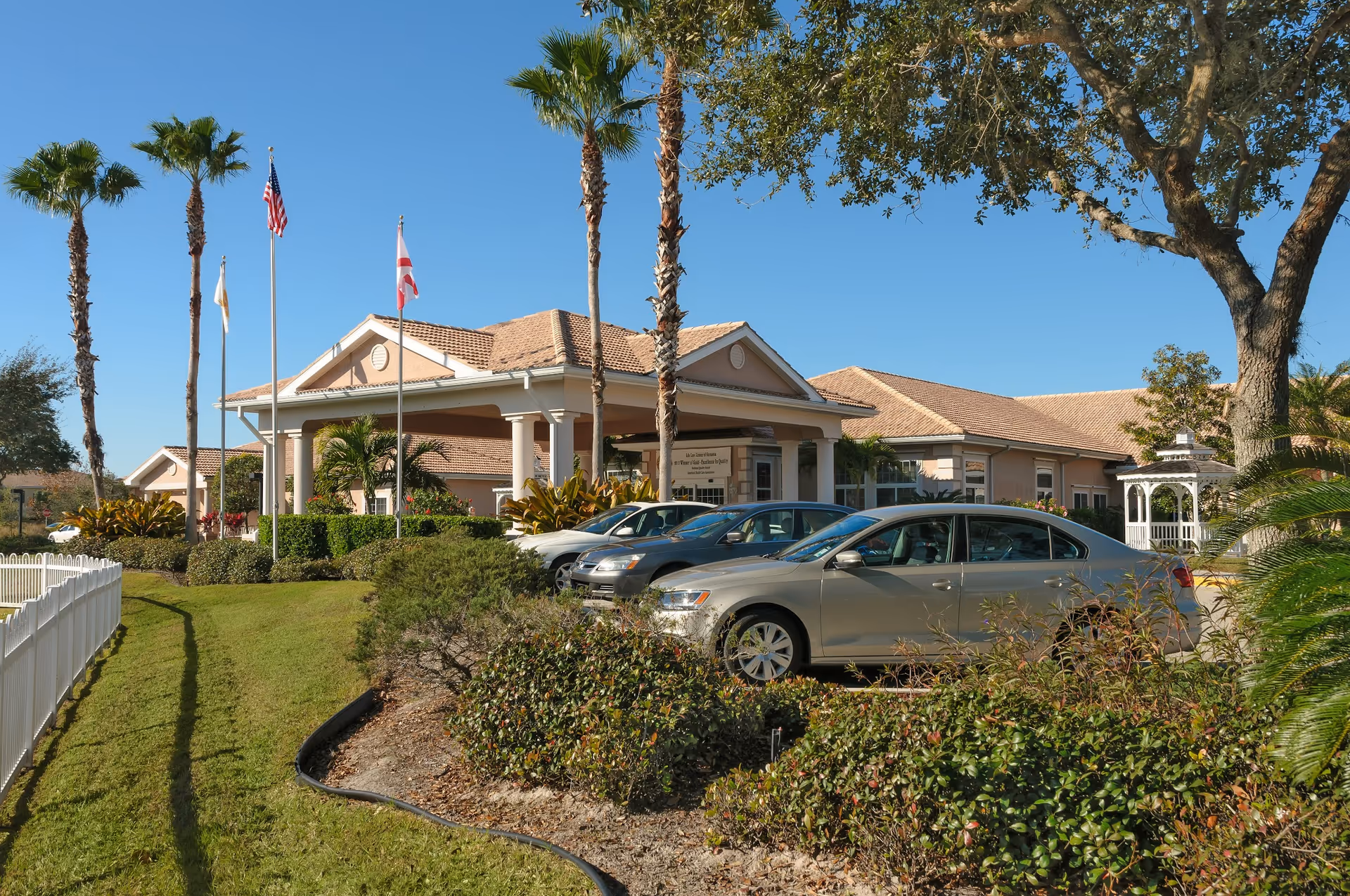 Front entrance of a single-story senior living facility with columns, flagpoles, palm trees, parked cars, and landscaped grounds.