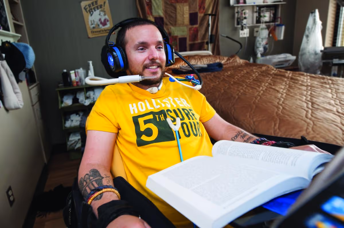 A man wearing a yellow Hollister t-shirt and headphones is sitting in a wheelchair in a bedroom. He has a device near his mouth to assist with communication and is reading a large open book. The room has a bed with a brown quilt, shelves with various items, and a poster on the wall that says 'FREE BEER!'.
