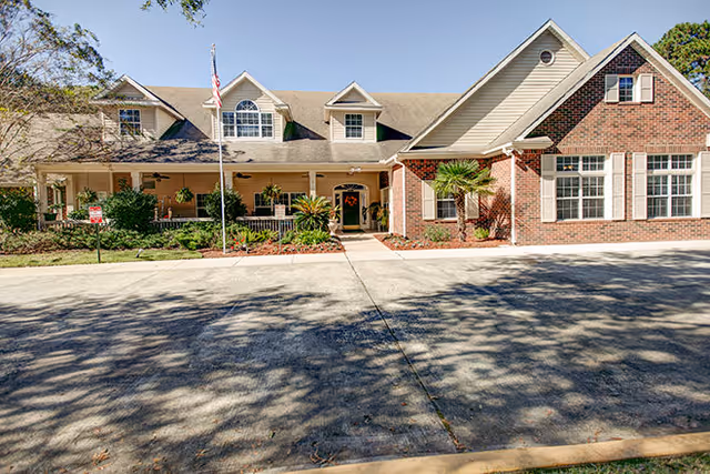 Front exterior view of a single-story assisted living facility building with a combination of brick and siding exterior, multiple windows, a covered porch with columns, an American flag on a flagpole, and landscaping including bushes and small trees.