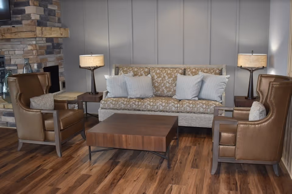 A cozy living room area with a floral patterned sofa adorned with light-colored cushions, two brown leather armchairs with matching pillows, a wooden coffee table, two side tables each holding a lamp, and a stone fireplace in the corner.