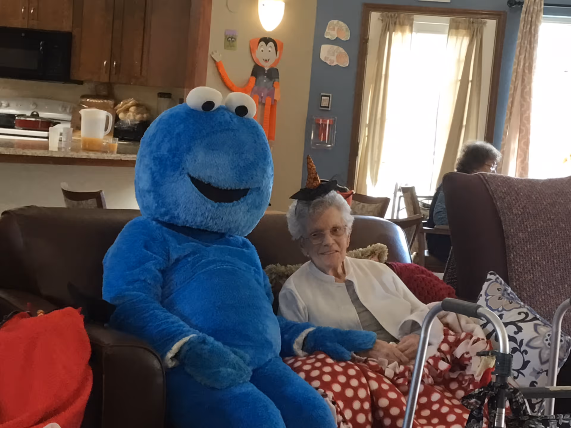An elderly woman wearing glasses and a small witch hat sits on a couch next to a person dressed in a large blue Cookie Monster costume. They are in a cozy living room area with a kitchen visible in the background. The elderly woman has a red and white polka dot blanket on her lap and a walker in front of her. Halloween decorations, including a paper vampire figure, are on the wall behind them.