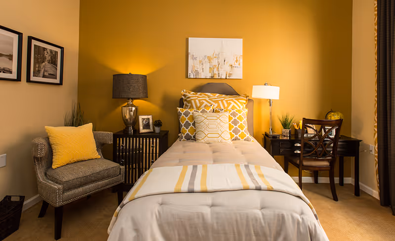 A cozy bedroom with a single bed centered against a mustard yellow wall. The bed is neatly made with a beige comforter and decorative pillows in yellow and white patterns. On the left side of the bed is a small nightstand with a silver lamp, a framed photo, and a small plant. Next to the nightstand is a gray armchair with a yellow pillow. On the right side of the bed is a dark wooden desk with a chair, a white lamp, a small plant, and a globe. Two black and white framed pictures hang on the adjacent wall, and a piece of artwork hangs above the bed.