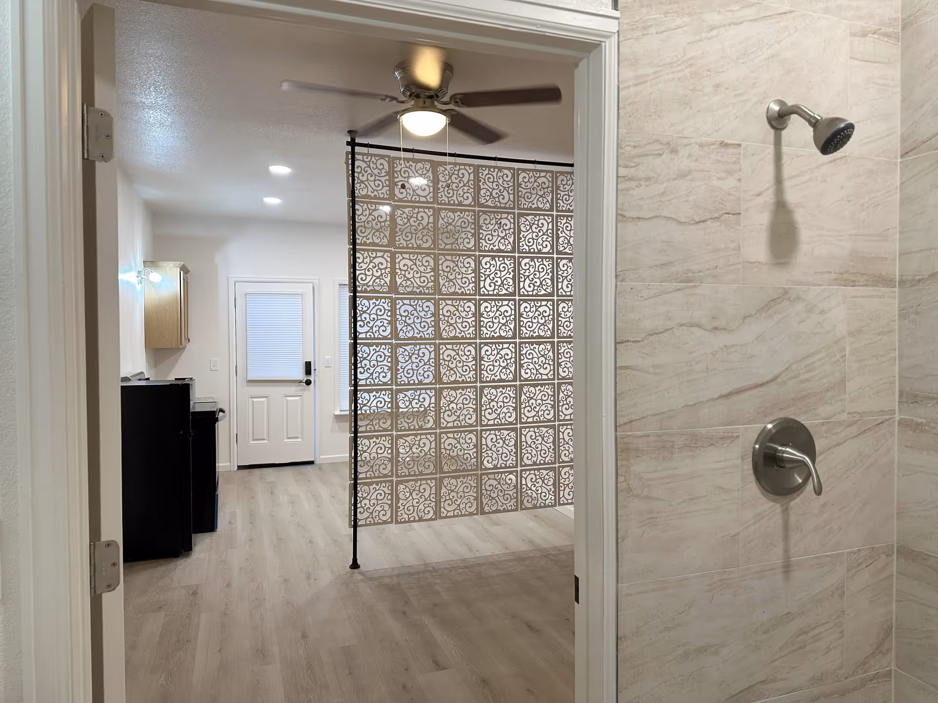 View from inside a tiled shower looking out into a room with light wood flooring, a decorative metal room divider, a ceiling fan with light, a door with a window, and a small kitchenette area with a black refrigerator and wooden cabinets.