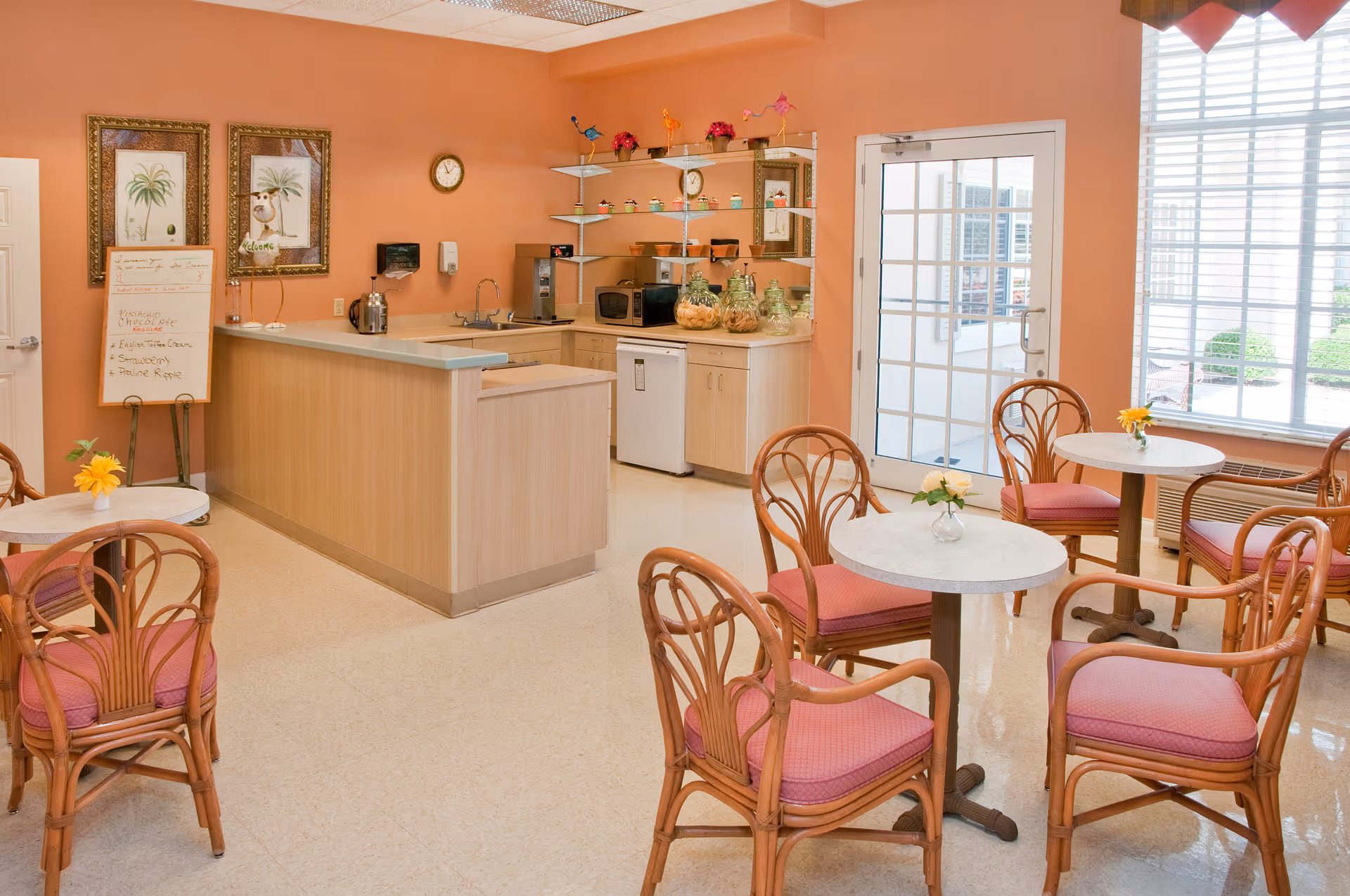 Bright communal dining area with round tables and wicker chairs facing a small kitchenette against a peach-colored wall.