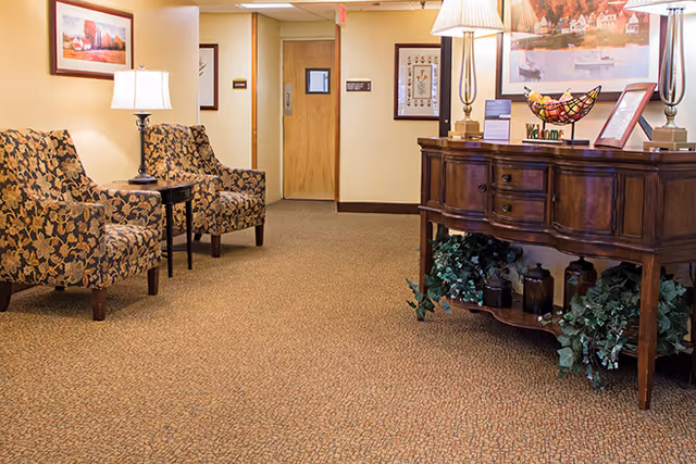 A cozy interior hallway area in a senior living facility with patterned armchairs, a small side table with a lamp, a wooden console table decorated with lamps, plants, and framed artwork on the walls.