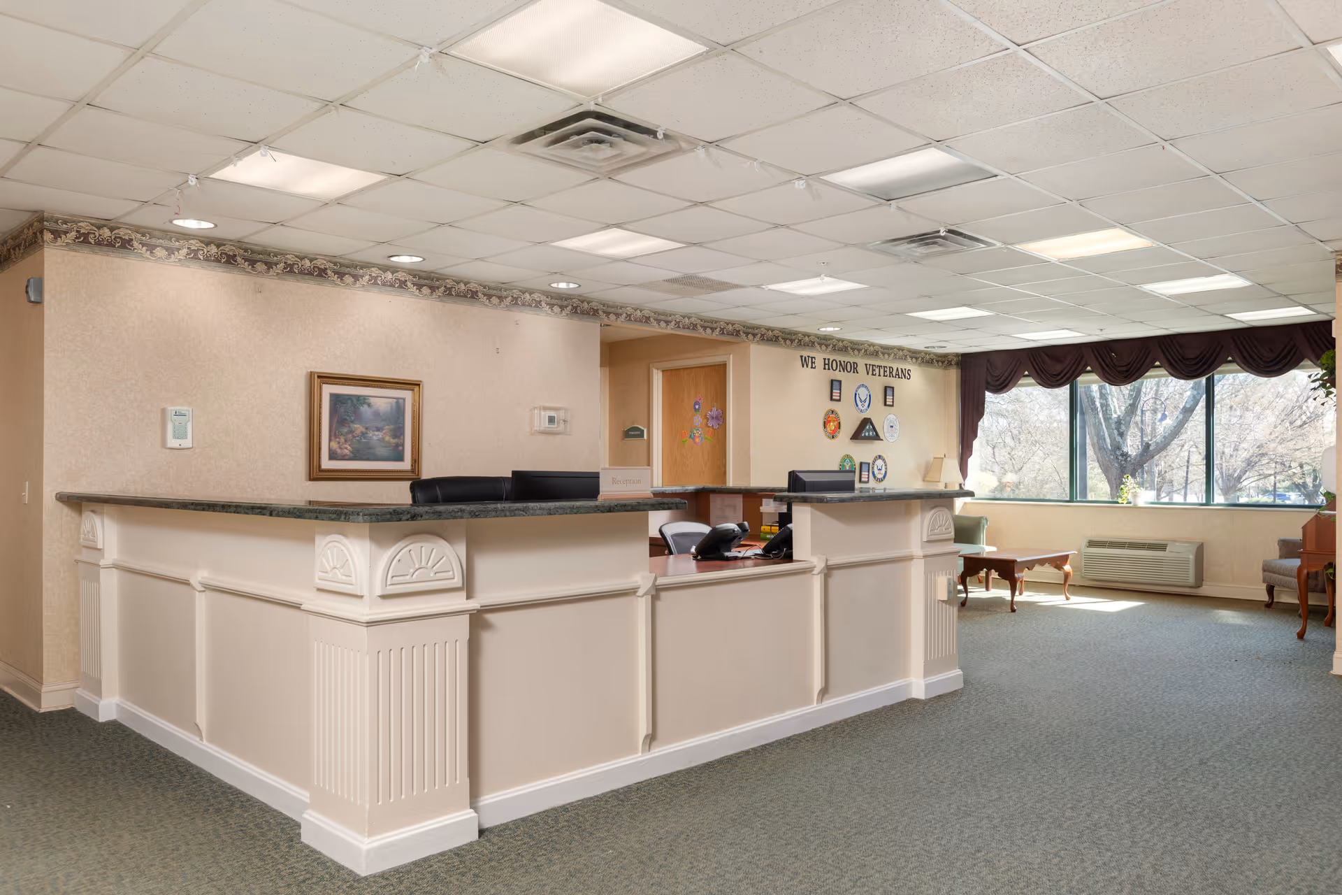 Reception area in a senior living facility with a large white reception desk, office chairs, a framed painting on the wall, and a wall display honoring veterans. The room has large windows with dark curtains, carpeted floor, and soft lighting from ceiling fixtures.