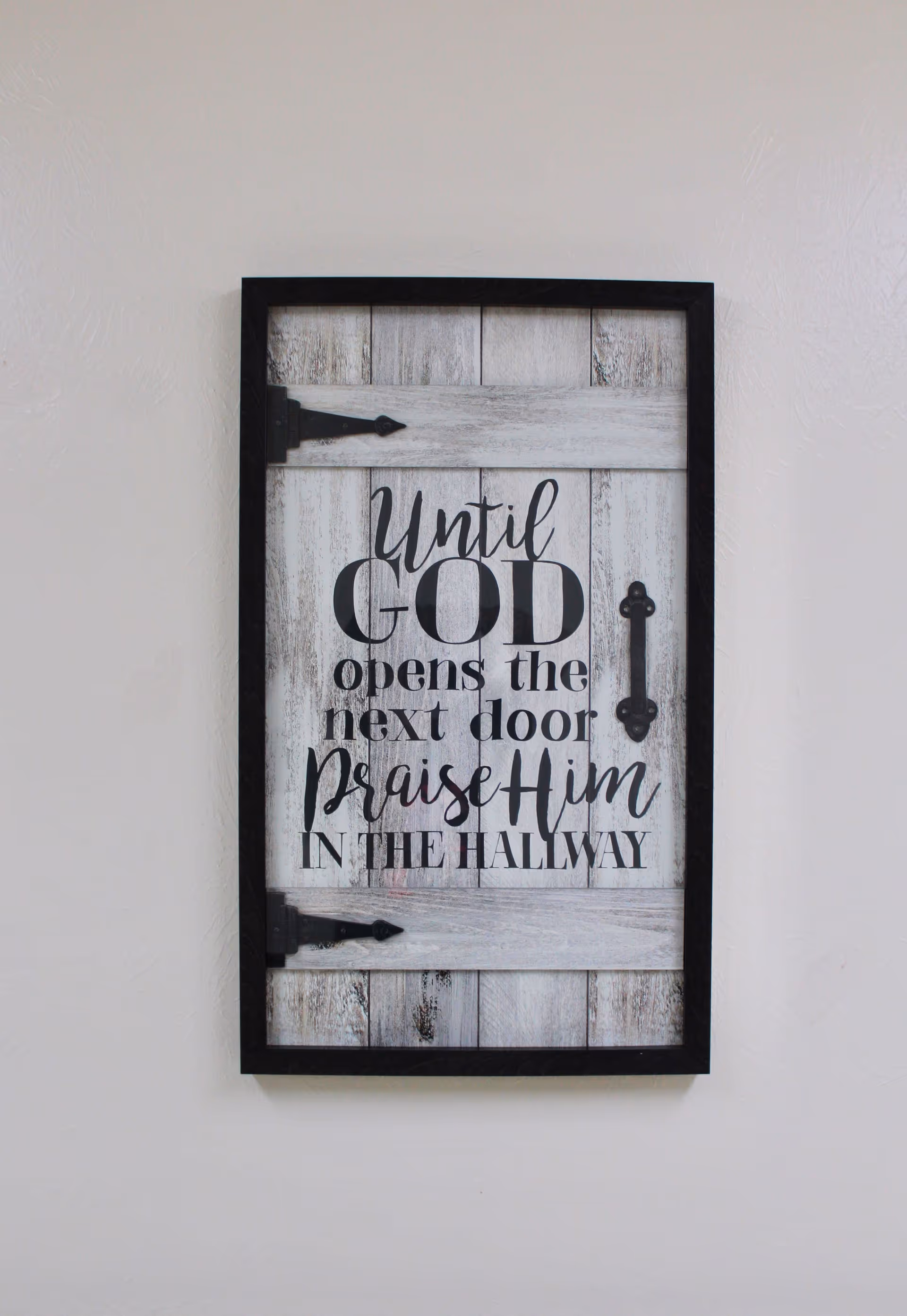 A decorative wooden wall sign designed to look like a rustic door with black hinges and handle. The sign reads: 'Until GOD opens the next door Praise Him IN THE HALLWAY'. The sign is mounted on a plain light-colored wall.