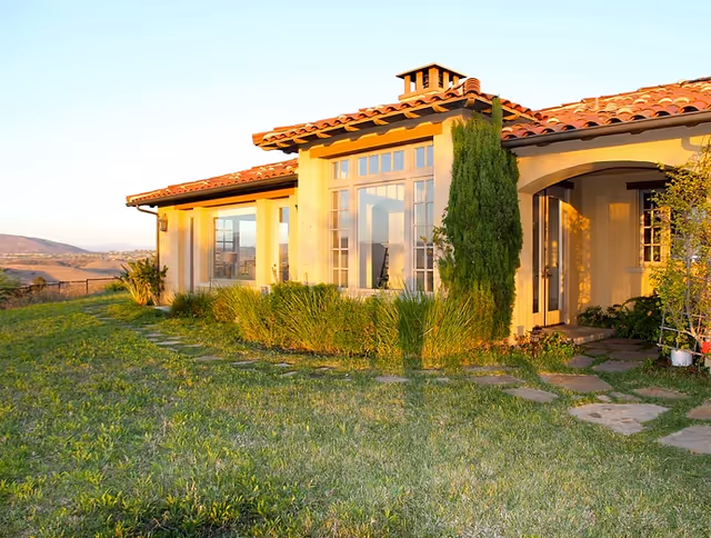 Sunlit stucco villa with red-tiled roof, large windows, and surrounding green lawn.