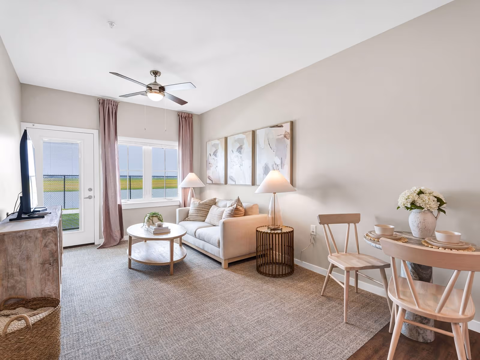 Bright, airy living room with a sofa, round coffee table, TV on a console, and a small dining table by a windowed door.