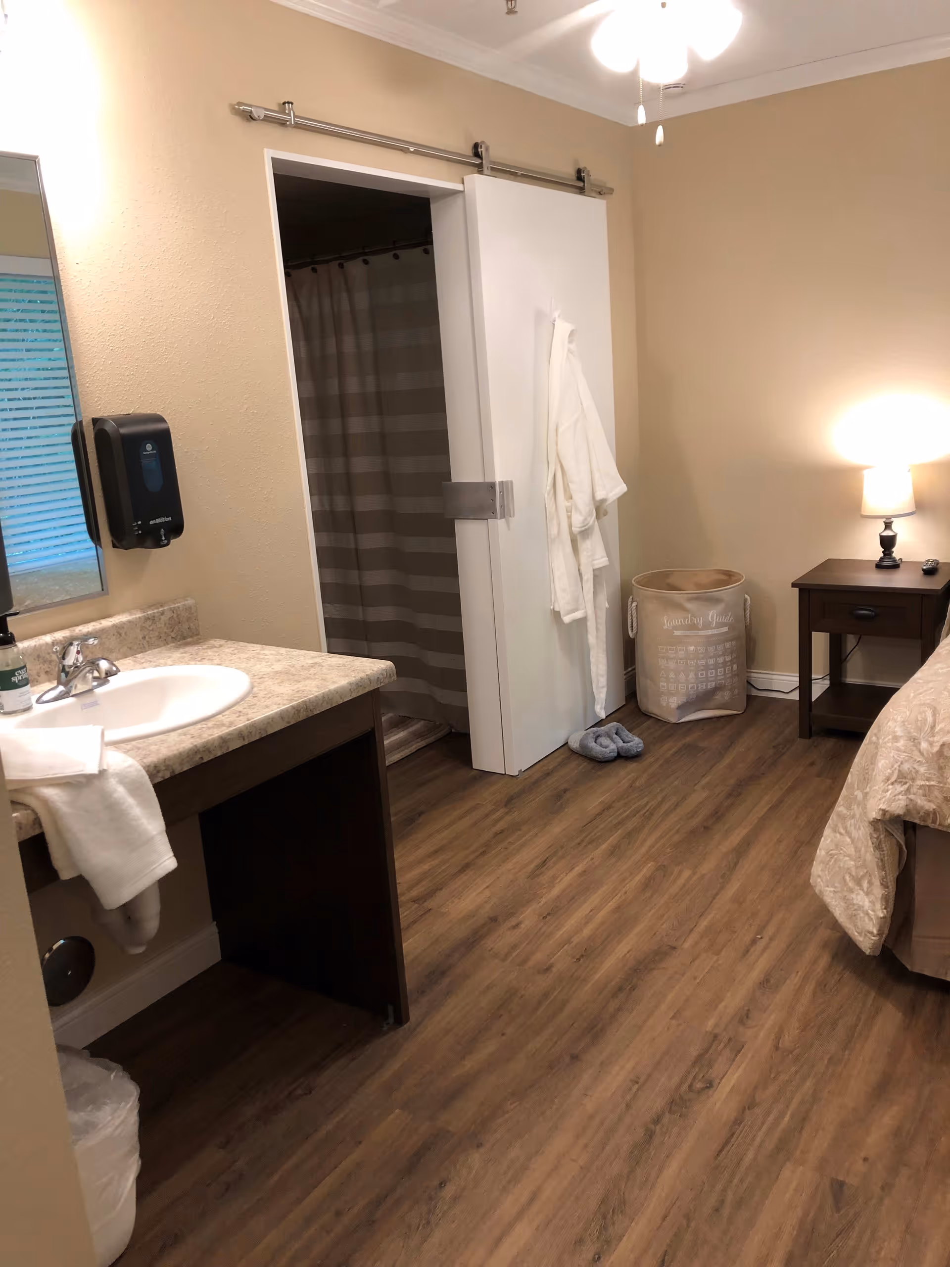 Interior view of a senior living facility room at Corridor Crossing Place featuring a sink with countertop and mirror on the left, a sliding door partially open revealing a shower with a striped curtain, a white robe hanging on the door, a laundry basket labeled 'Laundry Guide', a pair of slippers on the floor, a nightstand with a lamp and remote control, and part of a bed with beige bedding. The room has wood-style flooring and beige walls.
