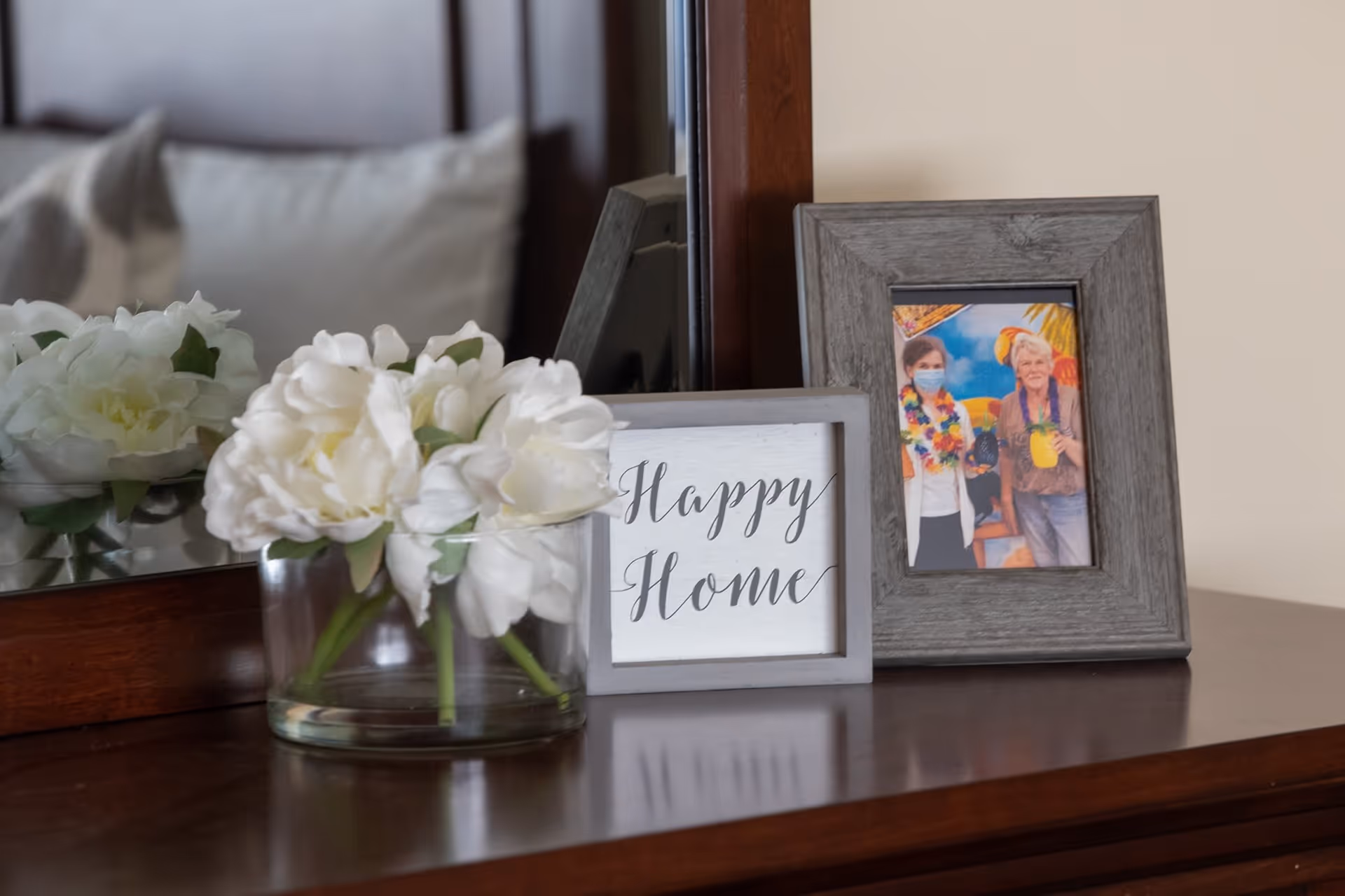 A wooden dresser top with a glass vase holding white flowers, a small framed sign that reads 'Happy Home', and a framed photo of two women, one wearing a face mask and colorful leis, standing in front of a tropical backdrop.