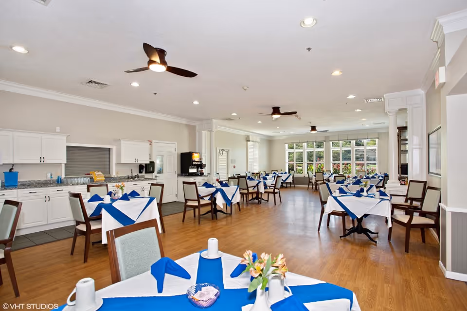 A spacious dining room with multiple tables covered in white tablecloths and blue napkins, each set with cups and small flower arrangements. The room has wooden flooring, white walls, ceiling fans, recessed lighting, and large windows letting in natural light. There is a counter with cabinets and a beverage dispenser along one wall.