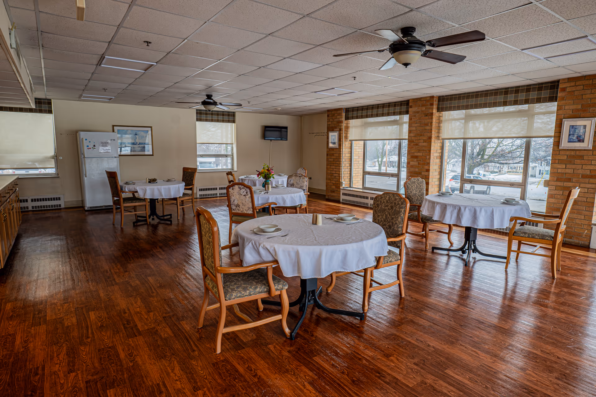 A dining room with several round tables covered with white tablecloths, each set with plates and bowls. The room has wooden floors, large windows with roller shades, brick walls, ceiling fans, and a refrigerator in the corner.
