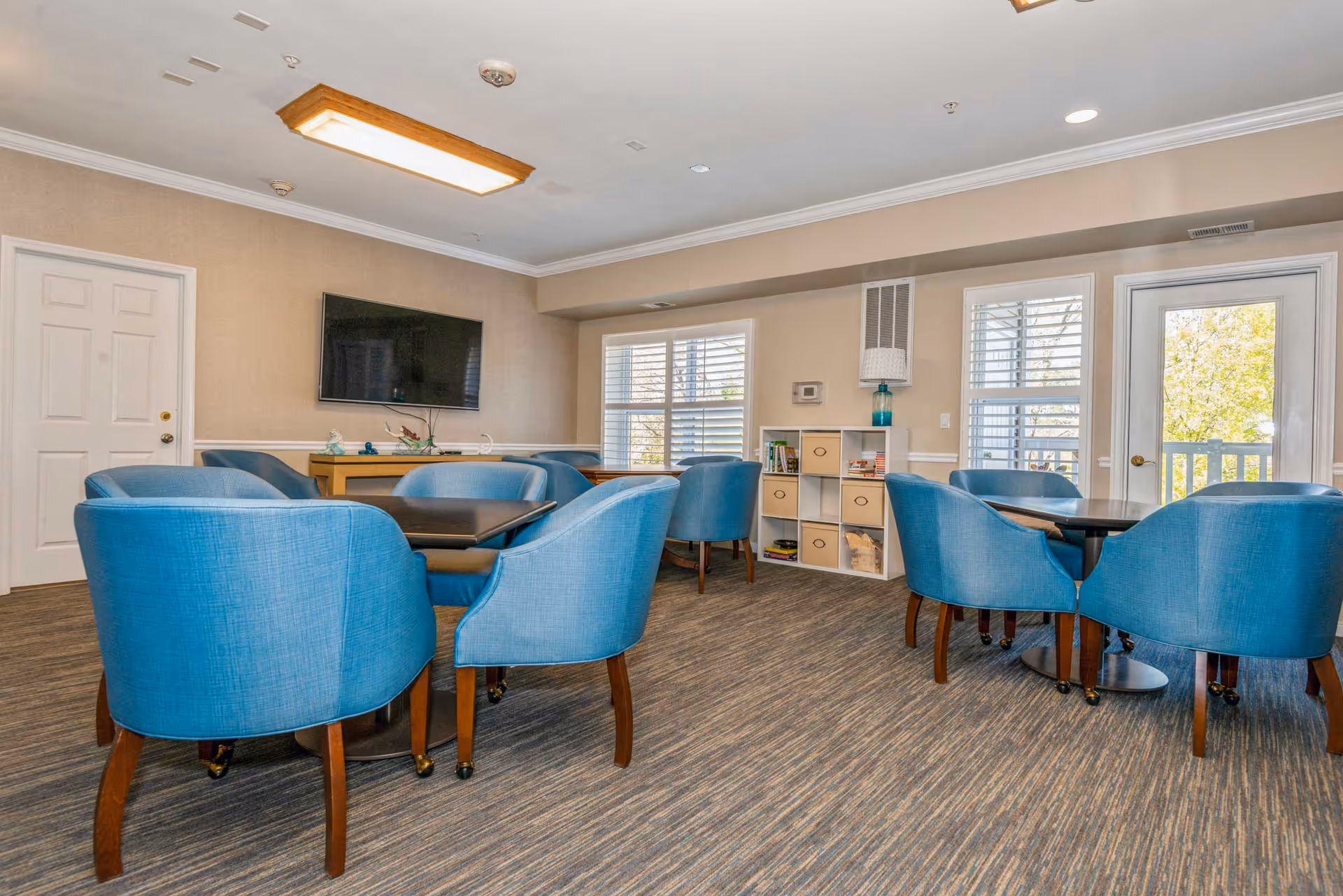 A bright and spacious common area with multiple blue upholstered chairs arranged around dark wooden tables. The room features beige walls, a large flat-screen TV mounted on the wall, windows with white blinds, a white door, and a white shelving unit with storage bins and decorative items. The floor is carpeted with a patterned design.