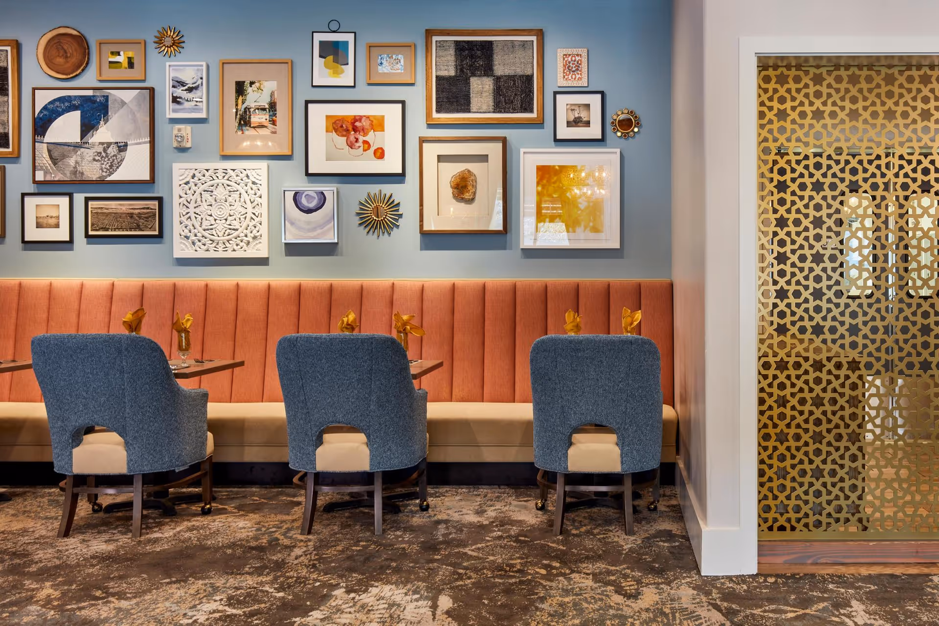 A stylish dining area with a long coral-colored upholstered bench against a blue wall adorned with various framed artworks and decorative pieces. In front of the bench are three tables, each paired with a blue upholstered chair. To the right, a decorative gold lattice partition partially separates the space.