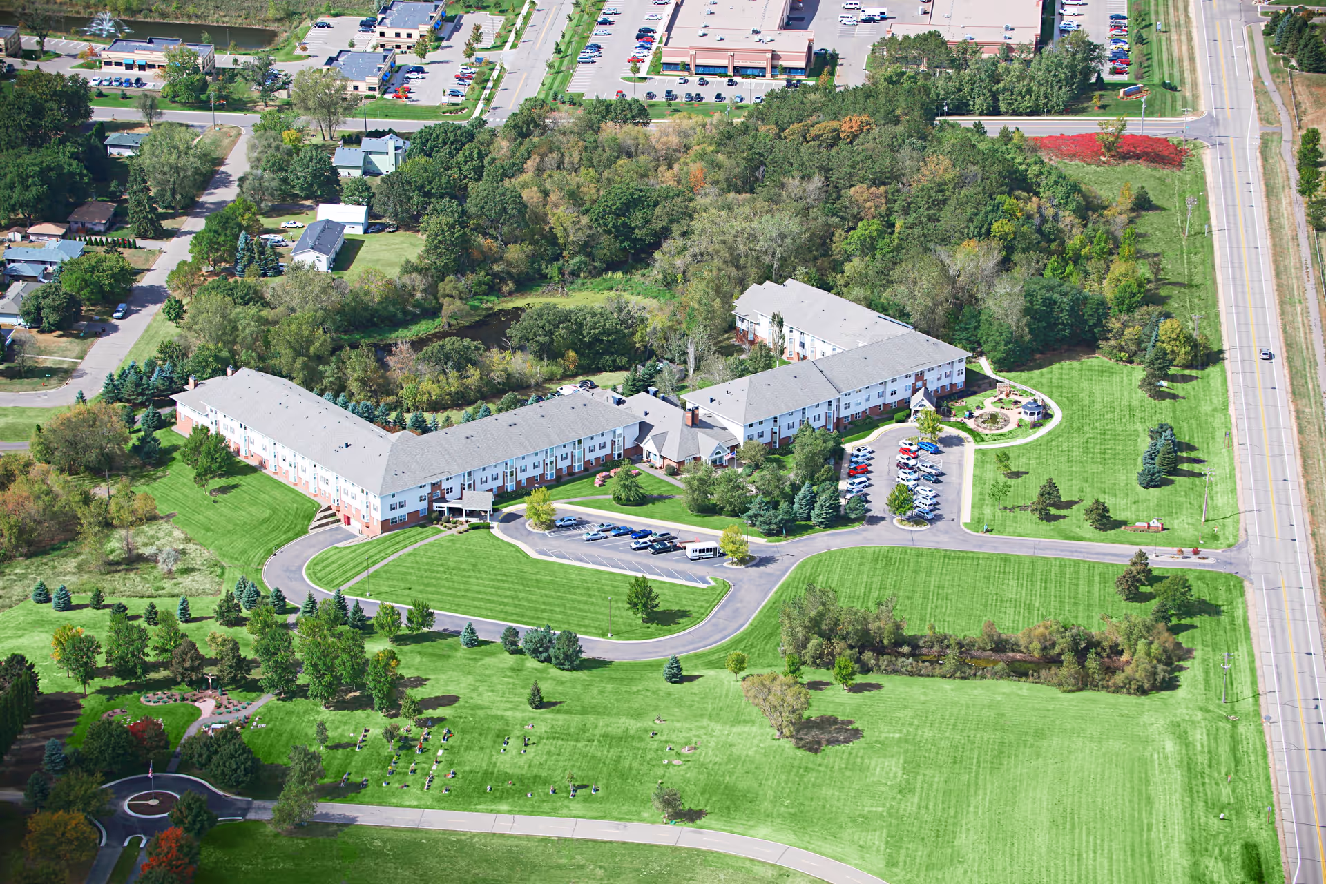 Aerial view of the Oak Meadows Senior Living complex showing an L-shaped building surrounded by lawns, trees, driveways and parking areas.