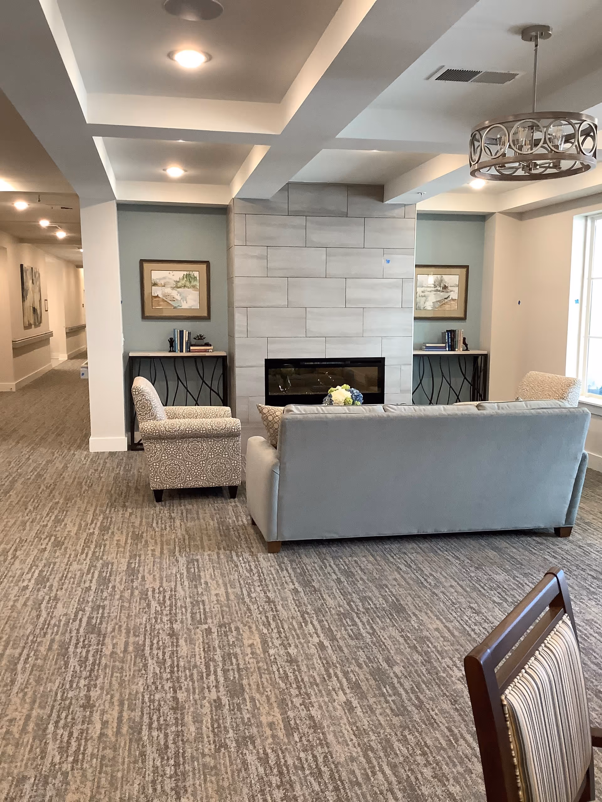 A cozy seating area in a senior living facility featuring a gray couch and two patterned armchairs arranged around a modern fireplace with a tiled surround. There are two small tables with books and decorative items on either side of the fireplace. The room has a carpeted floor, recessed ceiling lights, and a decorative hanging light fixture. A hallway extends to the left, and a window is visible on the right side.