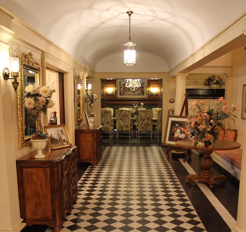 A long hallway with a black and white checkered floor leading to a dining area with floral upholstered chairs and a chandelier. The hallway has wooden furniture with floral arrangements and framed pictures, ornate gold-framed mirrors, and warm lighting fixtures on the walls. There is a round wooden table with a large floral arrangement on the right side and a cushioned bench with pillows.
