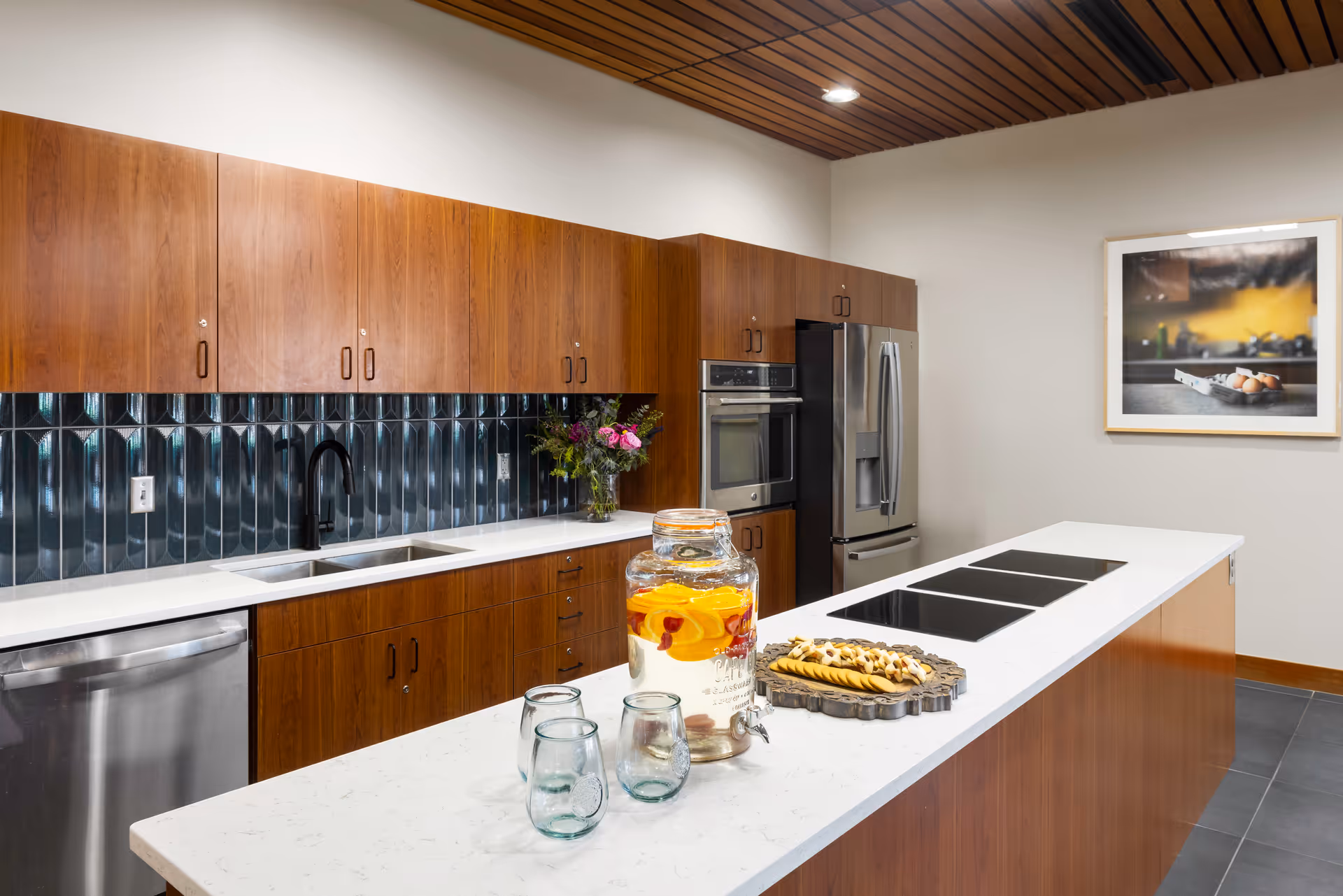 Modern kitchen with wooden cabinets, a white countertop island with three glass cups and a large glass beverage dispenser filled with water and fruit slices. The kitchen features a double sink, stainless steel dishwasher, built-in oven, and refrigerator. A framed picture of eggs on a kitchen counter hangs on the wall.