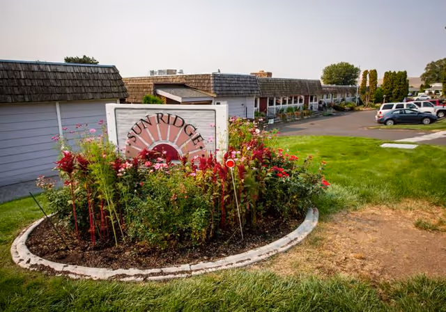 Outdoor view of Sun Ridge Retirement facility showing a flower bed with a sign that reads 'Sun Ridge' surrounded by various plants and flowers. In the background, there are single-story buildings with shingled roofs and a parking area with several cars.