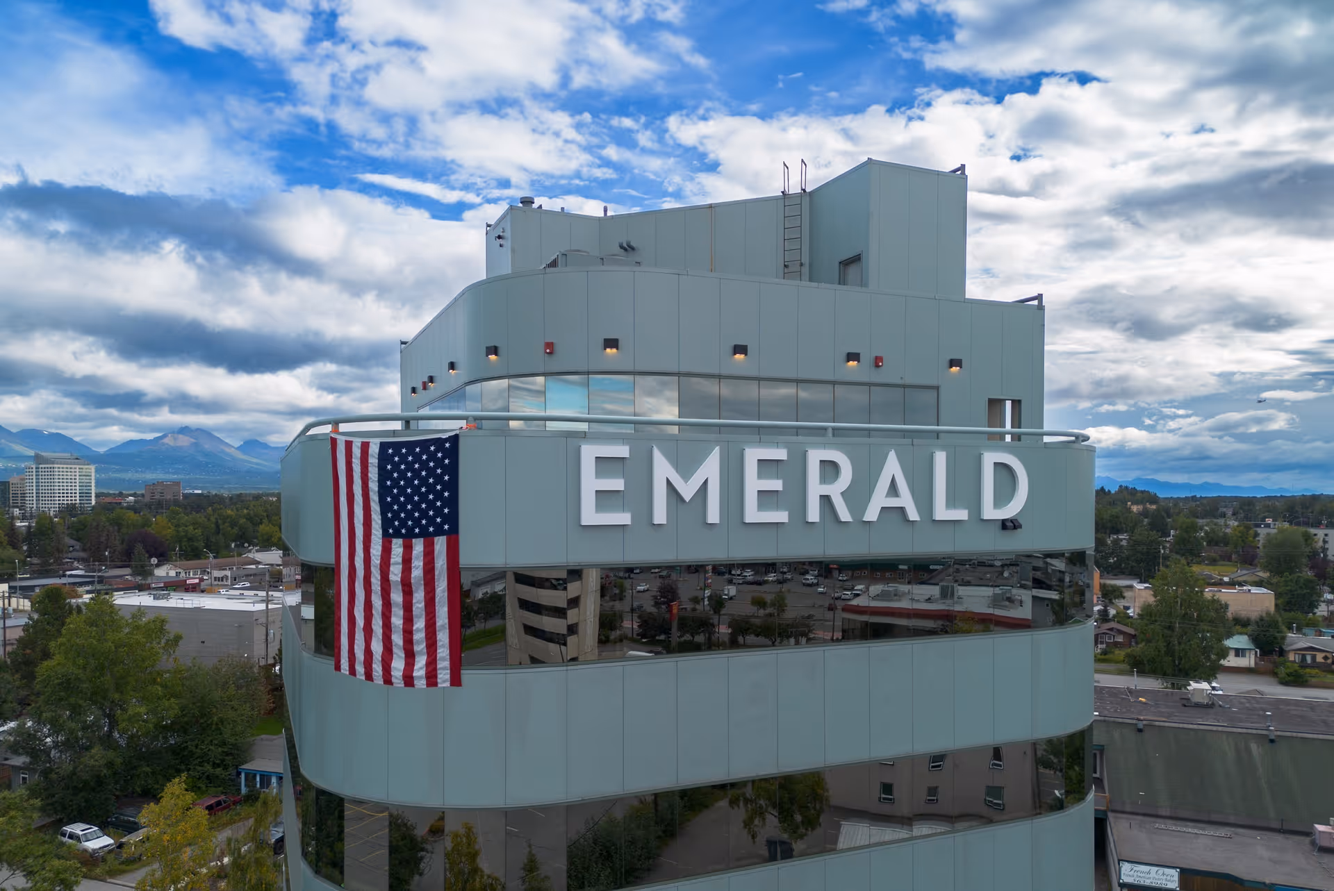 Exterior view of a modern multi-story building with large windows and the word 'EMERALD' prominently displayed on the facade. An American flag hangs vertically on the side of the building. The sky is partly cloudy with mountains visible in the background and a cityscape below.