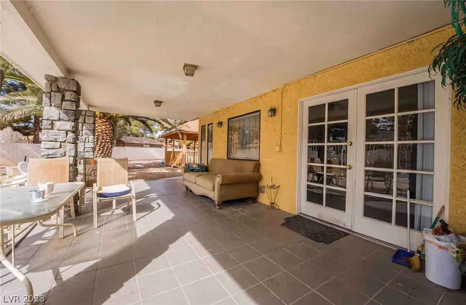 Covered outdoor patio area with tiled floor, stone pillars, a beige sofa against a yellow wall, glass double doors, and a table with chairs. There are palm trees and a wooden gazebo visible in the background.