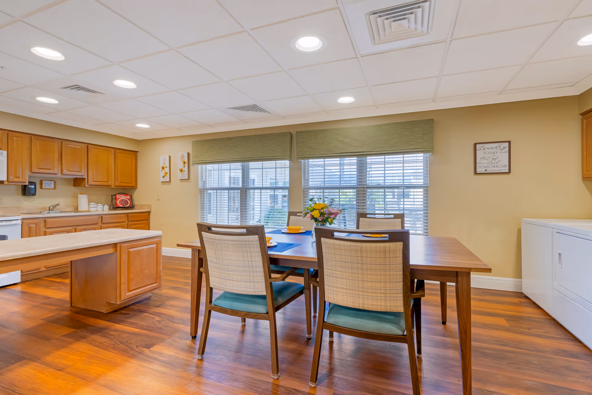 A bright kitchen and dining area with wooden floors and cabinetry. The dining table is set with four chairs and has a flower arrangement in the center. Large windows with green valances allow natural light to fill the room. On the right side, there is a white washer and dryer unit. The walls are painted beige and decorated with small framed art pieces.