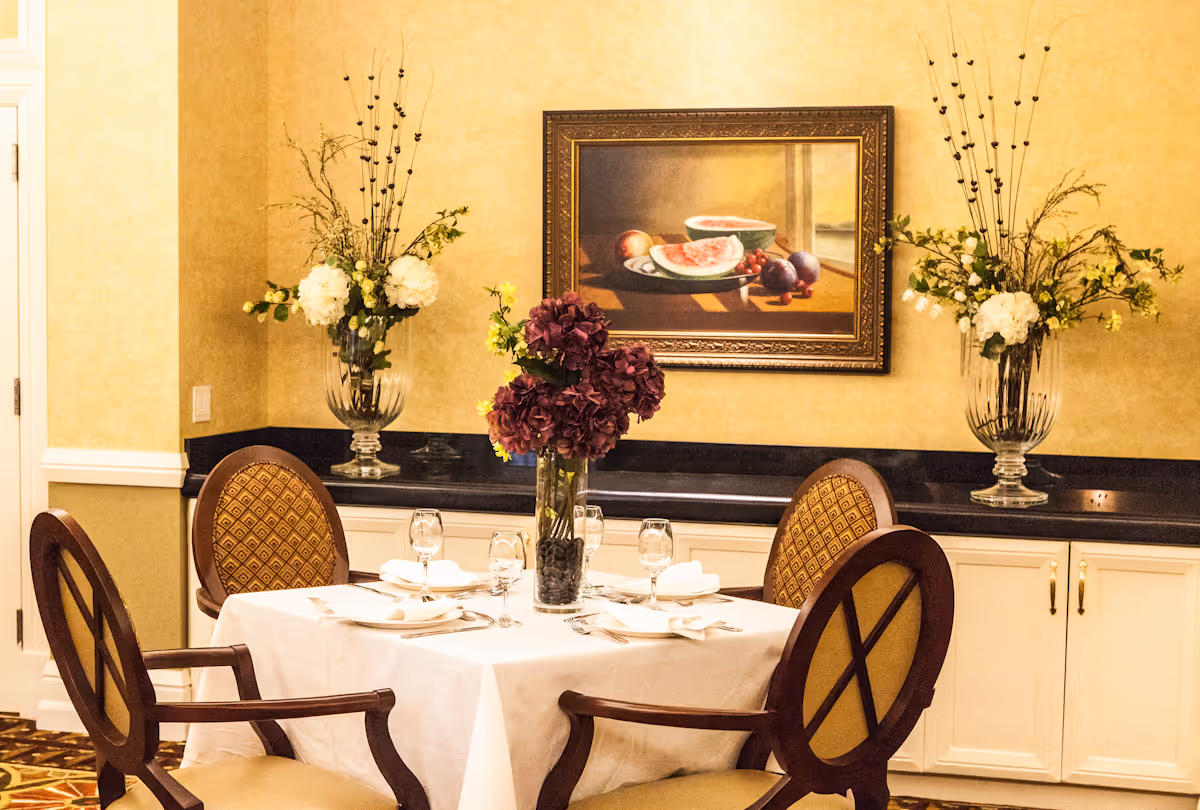 A dining room table set for four with white tablecloth, napkins, plates, and glasses. The table has a centerpiece of purple flowers in a glass vase. Behind the table is a black countertop with two large glass vases containing white flowers and decorative branches. A framed painting of fruit hangs on the beige wall above the countertop.